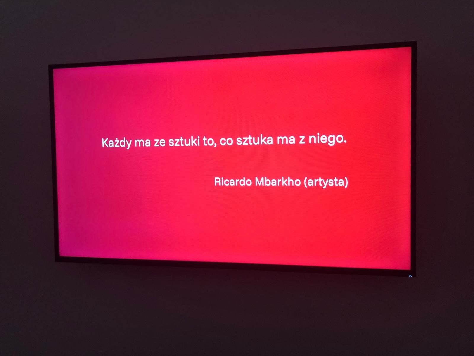 Écran affichant une citation de Ricardo Mbarkho en polonais : « Każdy ma ze sztuki to, co sztuka ma z niego. »